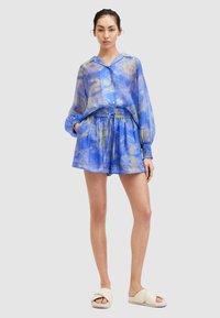 Svijetloplava košulja s dugim rukavima i žutim apstraktnim uzorcima, uparena s plavim šortsevima. Nosila se uz krem sandale na sklizak.