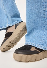Sneakers featuring a mix of black leather, beige suede, and textured beige rubber sole with ridges. Worn with light blue denim pants.