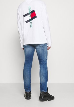 Persona vistiendo vaqueros azules descoloridos, deportivas negras y una camiseta blanca de manga larga con un logo de Tommy Jeans en rojo, negro y verde en la parte trasera.