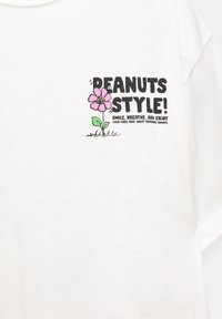 Hvid bomuld t-shirt med et farverigt grafik af en blomst og tekst, der siger "PEANUTS STYLE!" med yderligere sætninger i sort.