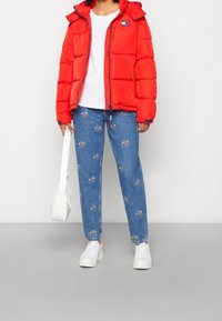 Red puffer jacket with a hood and zipper, white t-shirt, blue jeans featuring floral embroidery, and white sneakers. Holding a small white bag.