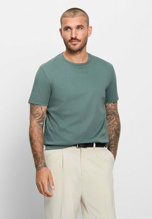 Uomo con capelli corti e tatuaggi sulle braccia, indossa una maglietta verde e pantaloni beige, in piedi davanti a uno sfondo bianco uniforme.