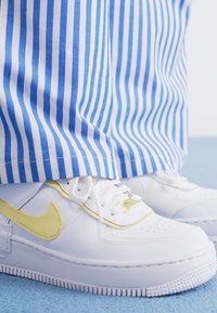 Bijele Nike tenisice s žutim naglasima i teksturiranom potplatom; u kombinaciji s plavim i bijelim prugastim hlačama. Pozadina je svijetloplave boje.