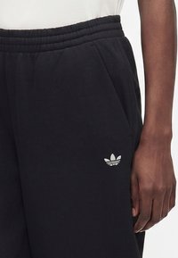 Άτομο που φοράει μαύρο παντελόνι φόρμας Adidas με ελαστική μέση και λογότυπο Trefoil κοντά στην τσέπη, το χέρι ακουμπάει στο πλάι.