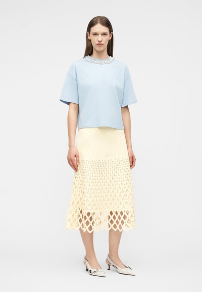 Femme portant un haut à manches courtes bleu clair avec un col décoratif, une jupe midi crème à motif en treillis, et des escarpins blancs à talons bas, debout immobile.