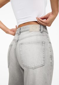 Persona che indossa jeans a vita alta grigi chiari e un top corto bianco, mostrando le tasche posteriori dei jeans e l'etichetta del marchio sulla vita.