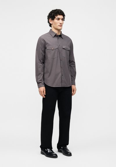 Άνδρας όρθιος, φορά γκρι πουκάμισο με κουμπιά και τσέπες στο στήθος, μαύρο παντελόνι και μαύρα επίσημα παπούτσια μπροστά σε απλό λευκό φόντο.