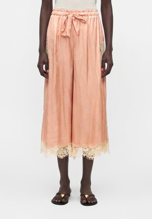 Μοντέλο που φοράει φαρδιά, κομμένα σατέν παντελόνια ροδακινί χρώματος με δαντέλα στα τελειώματα και πλαϊνές τσέπες, συνδυασμένα με μαύρα σανδάλια με χρυσές καρδιές.