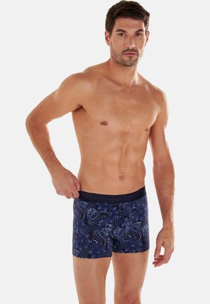 Oberkörperfreier Mann, der marineblaue Boxershorts mit Paisley-Muster richtet, stehend vor einem einfarbigen hellen Hintergrund.