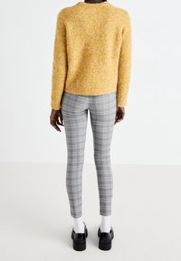 Texturerad gul tröja, åtsittande grå rutiga leggings, vita strumpor och svarta kraftiga skor, sedd bakifrån mot en ljus bakgrund.