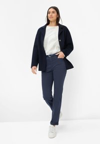Cappotto blu navy con colletto a revers, maglione leggero a trama sottile con motivo delicato e pantaloni blu navy. Scarpe da ginnastica bianche completano l'outfit.