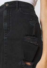 Černé džíny z denimu s několika roztrženými místy na stehně, nošené s zasunutým bílým topem a viditelným okrajem rukávu na pravé straně.