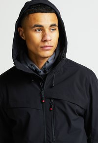 Černá vodotěsná bunda s kapucí, předním zipem, červenými detaily a stahovacími šňůrkami. Má hladkou texturu a prodyšnou vnitřní vrstvu.