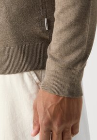 Hnědý pletený svetr s žebrovanými manžetami, vyrobený z texturované látky. Etiketa viditelná u švu, ruka položená na světlé tkanině.
