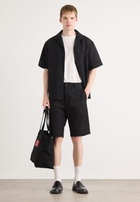 Cămașă neagră cu mânecă scurtă dintr-un material texturat, tricou alb dedesubt, pantaloni scurți negri și o geantă de tote neagră cu un eticheta roșie. Încălțări negre și șosete albe.