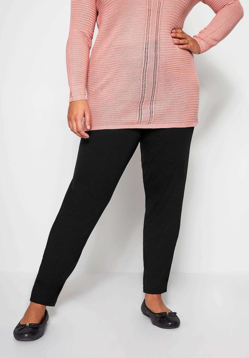 Mustat stretch-housut, joissa on kapea malli, yhdistettynä vaaleanpunaiseen neuletunikoihin, joissa on vaakasuoria raitoja ja pystysuoraa avointa ompelusta.