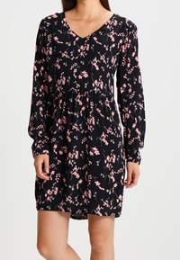 Robe noire à manches longues et col en V, ornée d'un motif floral en rose et gris. Le tissu est lisse avec une silhouette fluide.
