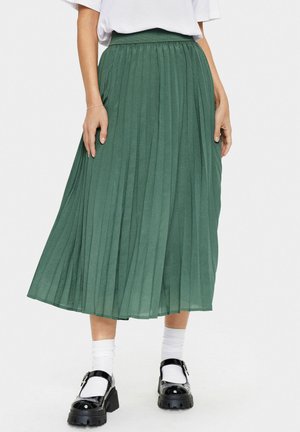 Gonna midi plissè verde realizzata in tessuto leggero, con una vita alta e un design fluido. Indossata con calzini bianchi e scarpe nere.