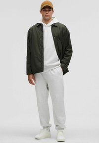 Herreoutfit bestående af en olivengrøn jakke, grå hættetrøje og matchende sweatpants, suppleret med hvide sneakers og en tan kasket.