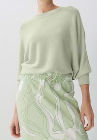 Hellgrüner Strickpullover mit lässiger Passform und geripptem Rundhalsausschnitt, kombiniert mit einem Rock, der ein wellenförmiges, abstraktes Muster in Grün und Weiß aufweist.