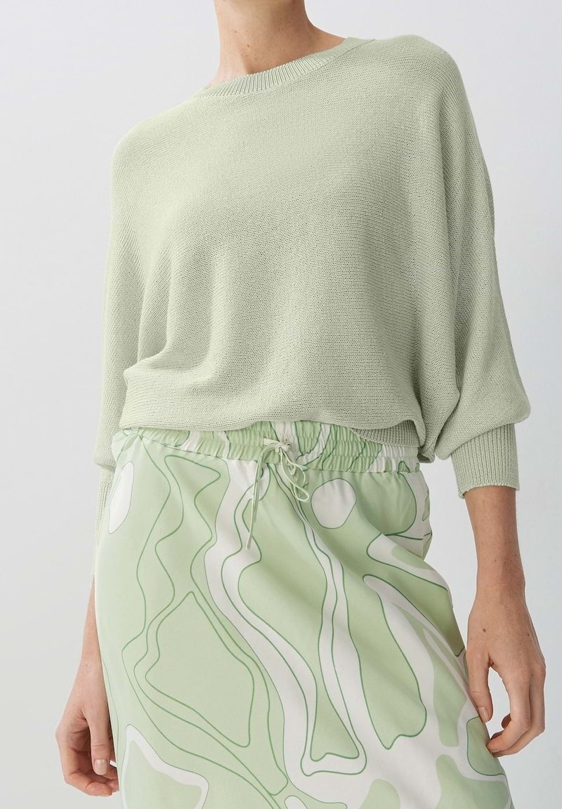 Hellgrüner Strickpullover mit lässiger Passform und geripptem Rundhalsausschnitt, kombiniert mit einem Rock, der ein wellenförmiges, abstraktes Muster in Grün und Weiß aufweist.