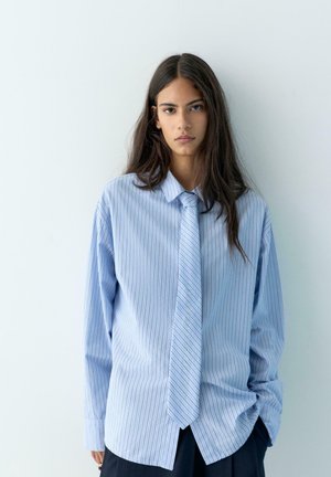 Młoda osoba z długimi ciemnymi włosami, ubrana w luźną niebieską koszulę w paski oraz pasujący krawat, stojąca na jednolitym jasnym tle.