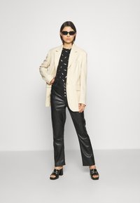 Um blazer oversized bege, uma blusa preta com padrão, calças de couro pretas de cintura alta e sandálias pretas grossas completam este conjunto moderno.
