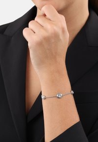 Pulsera de plata con tres piedras preciosas redondas y transparentes engastadas en una delicada cadena. Llevada en una muñeca, con un blazer oscuro visible en el fondo.