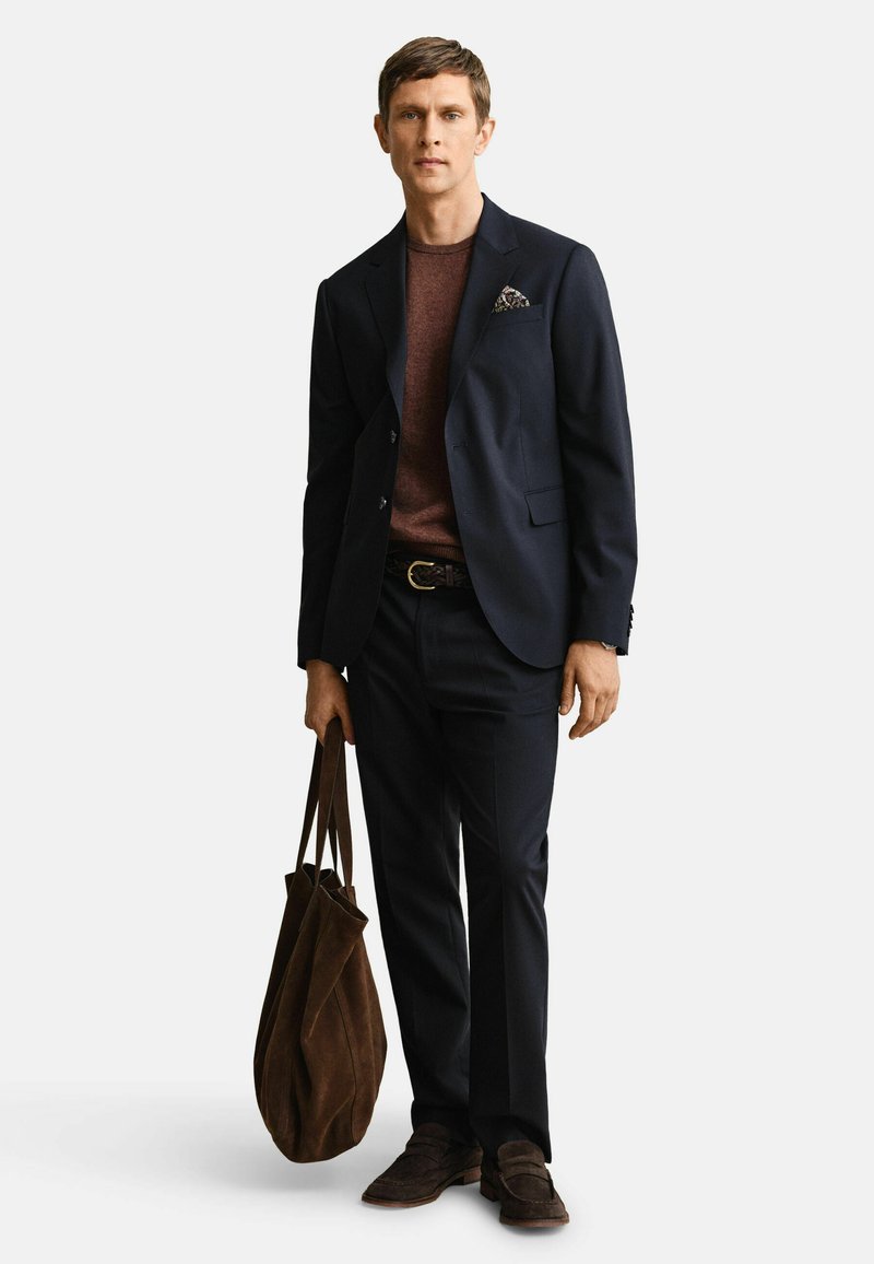 Homme portant un costume bleu marine avec un pull marron et des mocassins, tenant un grand sac fourre-tout en daim marron sur un fond uni.