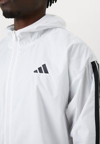 Casaco leve branco com capuz, apresentando um design de três riscas pretas ao longo das mangas e um logo preto no peito. Textura suave.