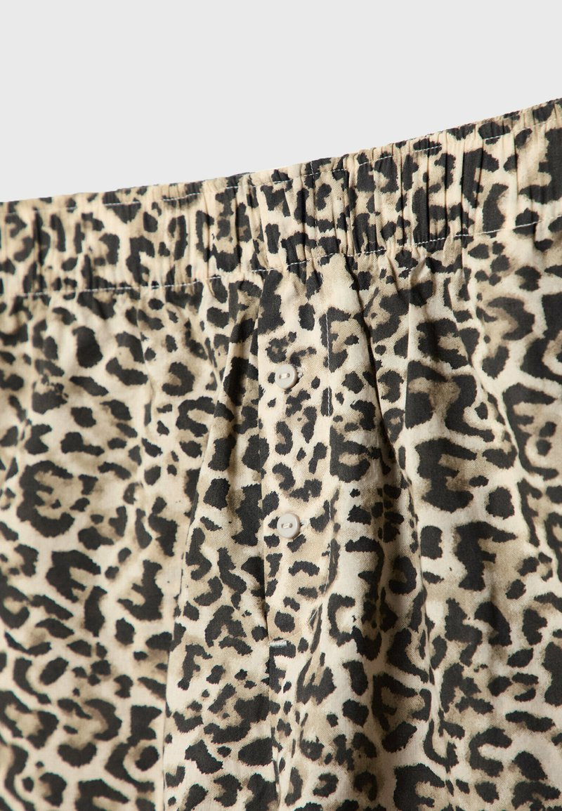 Leopardenmusterstoff mit einem hellbeigen Hintergrund. Verfügt über einen elastischen Bund und zwei weiße Knöpfe auf der Vorderseite. Strukturiert.