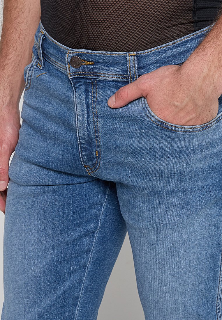 Osoba w niebieskich dżinsach z dopasowaną czarną siateczkową bluzką, z ręką spoczywającą w kieszeni dżinsów, na jednolitym tle.