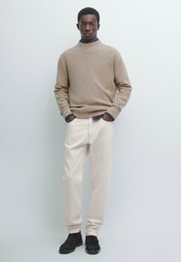 Beżowy sweter na drutach z wysokim kołnierzem, jasnokremowe spodnie oraz czarne teksturowane buty. Model stoi z rękami w kieszeniach na jednolitym tle.