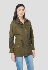 Olijfgroene jas met geborduurde bloemenpatronen, ritssluiting en drukknoopsluiting, twee borstzakken, taille met trekkoord en opstaande kraag.