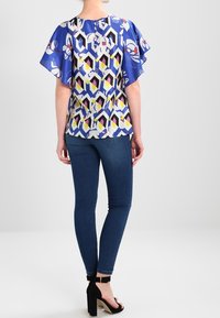 Blå mønstret bluse med korte, flagrende ærmer; geometrisk design i hvid, gul og pink; parret med mørke jeans og sorte højhælede sko.