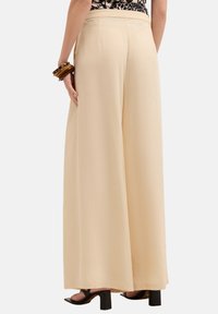 Pantaloni a gamba larga in un morbido tessuto beige, caratterizzati da una vita alta, una superficie liscia e una silhouette fluida. Scarpe nere con tacco a blocco visibili.