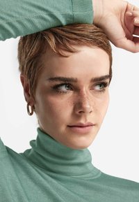 Długi rękaw, jasnozielony golf wykonany z miękkiej, teksturowanej tkaniny. Modelka ma krótkie włosy, widoczne piegi oraz kolczyki w kształcie kółka.