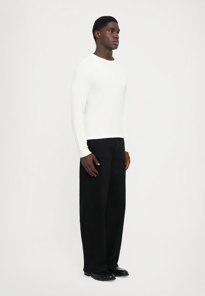 Un homme se tient face à droite, portant une chemise blanche à manches longues, un pantalon noir ample et des chaussures noires, devant un fond blanc uni.