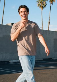 Nuori mies beigenvärisessä t-paidassa ja vaaleissa farkuissa hymyilee ja kävelee aurinkoisella ulkoilmaparkkipaikalla, taustalla palmuja.