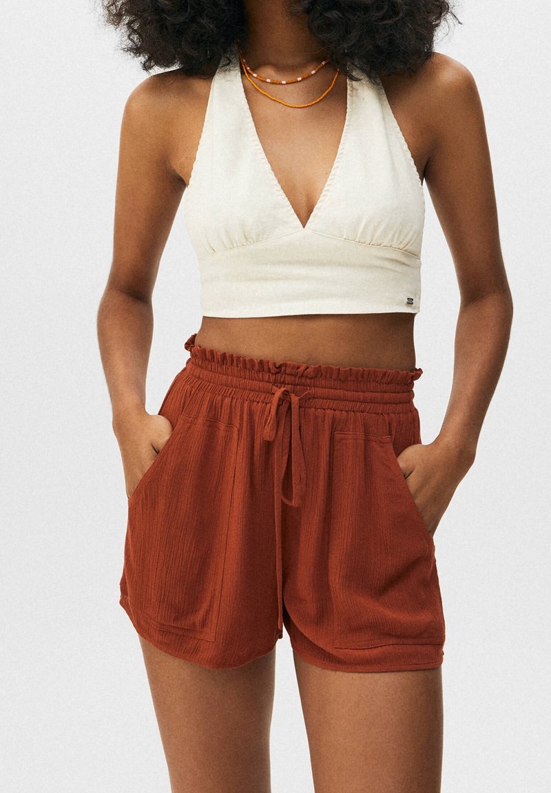 Hellcremiger Neckholder-Cropped-Top mit gerafften Details, kombiniert mit rostfarbenen, strukturierten Shorts mit elastischem Bund und Seitentaschen.