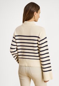 Stripete genser i krem og marineblå, med høy krage, ribbet kant og vevd tekstur. Lange, vide ermer med avslappet passform.