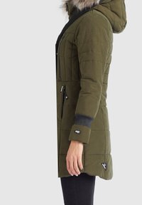 Grön vadderad jacka med huva, med svarta detaljer, ribbade muddarna och flera fickor. Inkluderar en pälsdetalj vid huvan.