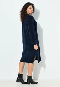 Navy gebreide jurk met lange mouwen, textuurstof, midden-kuit lengte, met zijsplitten; gecombineerd met zwarte enkellaarsjes met gouden accenten.