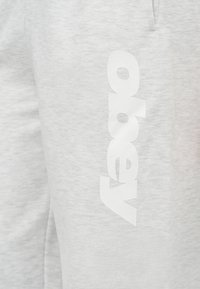 Ανοιχτό γκρι ύφασμα με κάθετο λευκό λογότυπο "Arena" τυπωμένο πάνω του, δείχνοντας λεπτομέρειες υφής και ραφής.