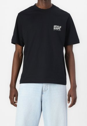 Man draagt een zwart T-shirt met witte tekst "Article Dept." op de borst en lichtblauwe spijkerbroek, staand tegen een witte achtergrond.