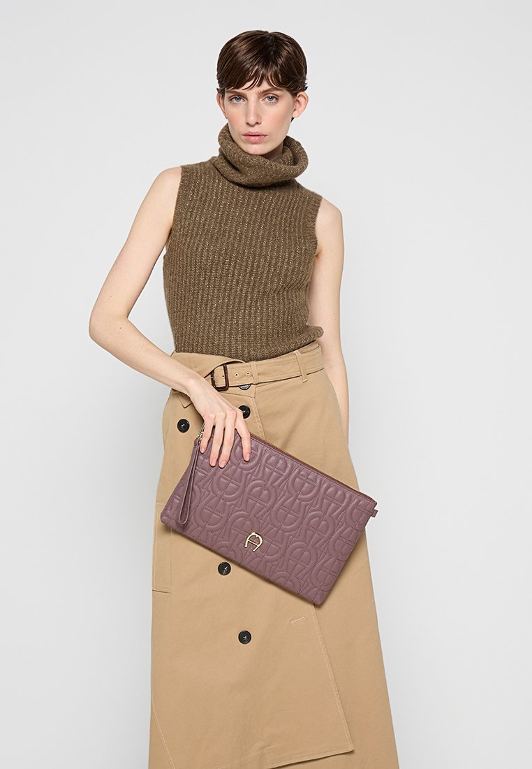 Brązowy sweter z golfem, beżowa spódnica z guzikami oraz prostokątna fioletowa torebka teksturowana z złotym logo i zamkiem.
