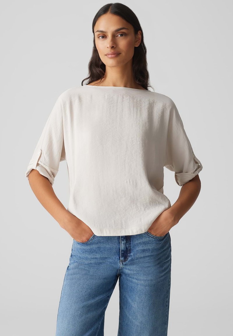 Helle Cremebluse mit geradem Schnitt und hochgekrempelten Ärmeln, aus weichem Stoff. Kombiniert mit blauen Jeans. Einfaches, klares Design.