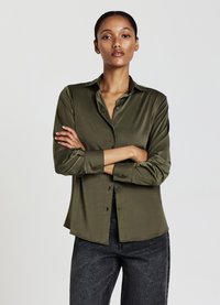 Camicia in satin verde oliva con chiusura a bottoni, colletto a punta e maniche lunghe, abbinata a jeans scuri. Tessuto liscio e vestibilità comoda.