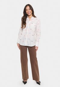 Camicia bianca con testo "Smile" in blu, rosa e rosso. Maniche lunghe, design a bottoni, abbinata a pantaloni marroni e scarpe nere.