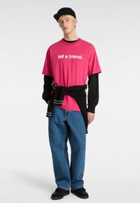 Lyserød T-shirt med hvid tekst, der siger "fortæl en ven", lagd oven på en sort langærmet skjorte. Sæt det sammen med løse blå jeans og sorte sko.